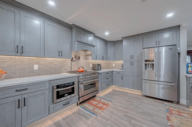 kitchen with gray cabinetry, appliances with stainless steel finishes, custom range hood, light stone countertops, and backsplash