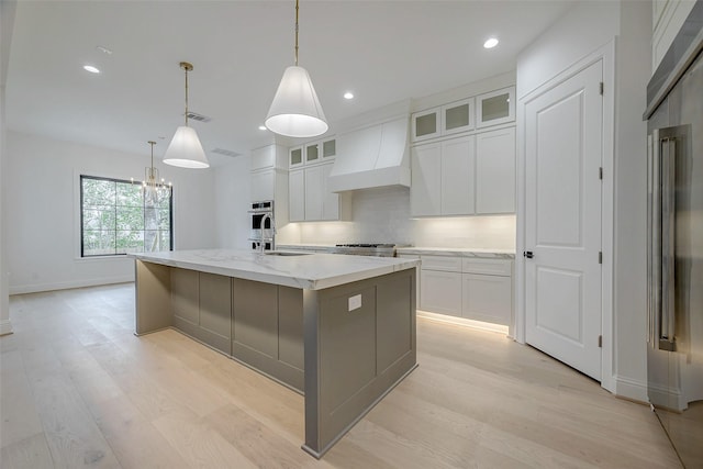 kitchen with premium range hood, decorative light fixtures, a center island with sink, light stone countertops, and white cabinets