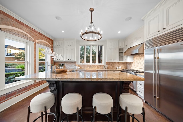 kitchen with stainless steel built in refrigerator, white cabinets, hanging light fixtures, exhaust hood, and range