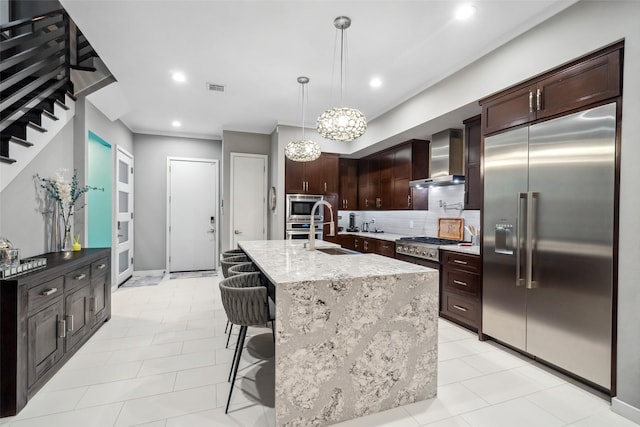 kitchen featuring an island with sink, a kitchen bar, built in appliances, light stone counters, and wall chimney range hood