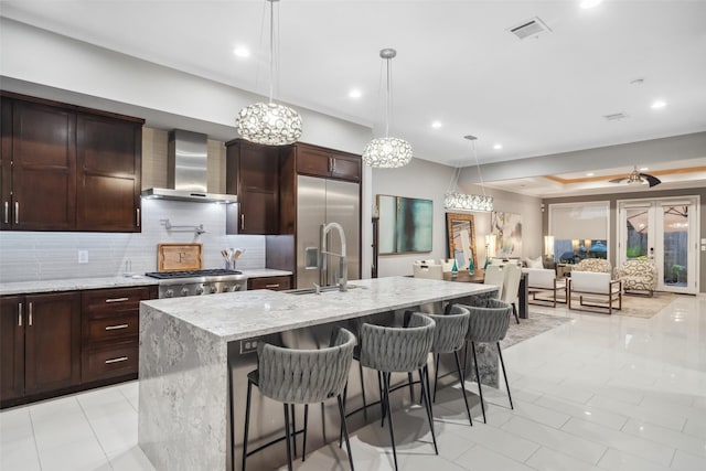 kitchen featuring appliances with stainless steel finishes, sink, light stone counters, a center island with sink, and wall chimney exhaust hood