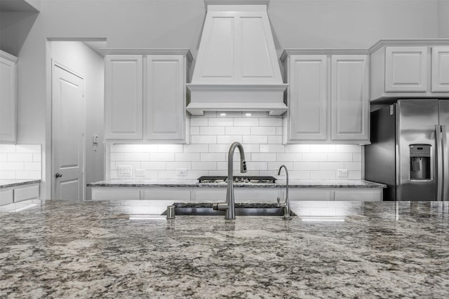 kitchen with custom exhaust hood, white cabinetry, tasteful backsplash, stainless steel fridge, and light stone countertops