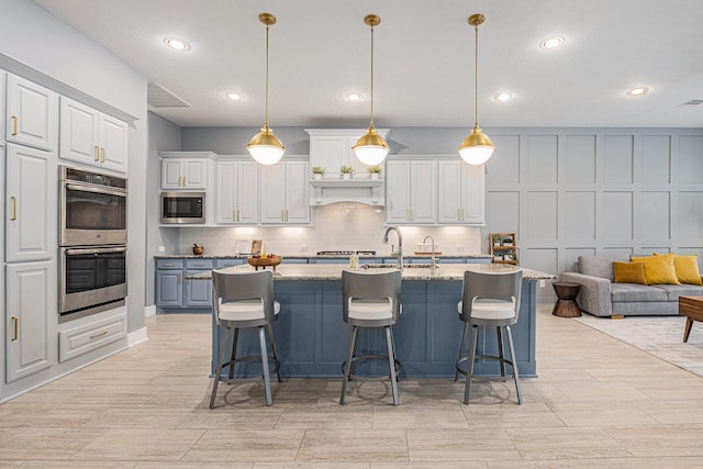 kitchen with light stone counters, built in microwave, stainless steel double oven, hanging light fixtures, and a kitchen island with sink