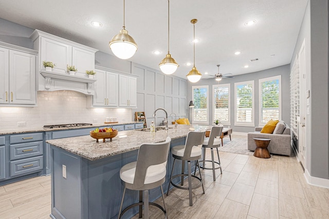 kitchen with an island with sink, hanging light fixtures, sink, light stone counters, and white cabinets