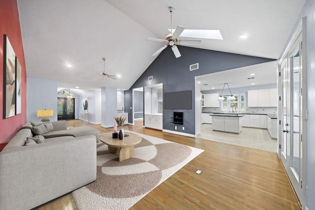 living room with high vaulted ceiling, a skylight, sink, ceiling fan, and light wood-type flooring