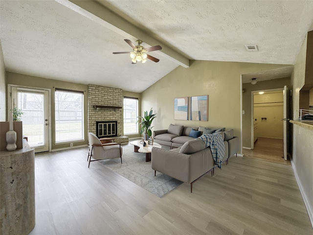 living room with lofted ceiling with beams, ceiling fan, a brick fireplace, a textured ceiling, and light hardwood / wood-style flooring