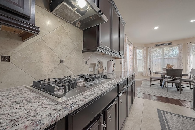 kitchen with wall chimney range hood, backsplash, light stone counters, stainless steel gas cooktop, and light tile patterned flooring