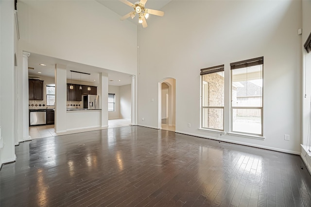 unfurnished living room featuring baseboards, arched walkways, a towering ceiling, ceiling fan, and dark wood-type flooring