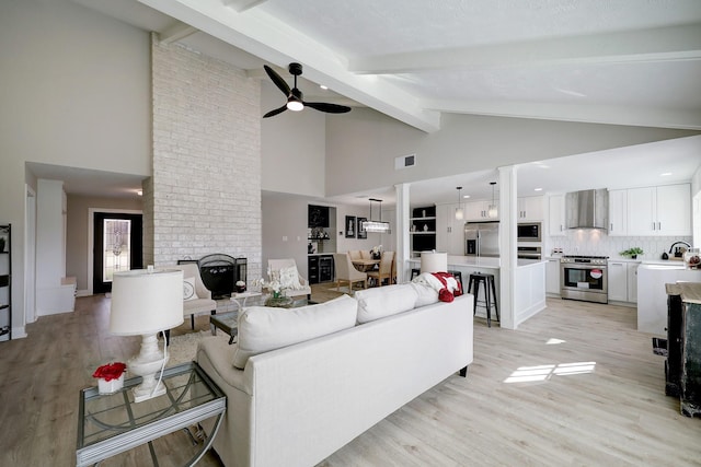 living room with ceiling fan, beam ceiling, a brick fireplace, and light wood-type flooring