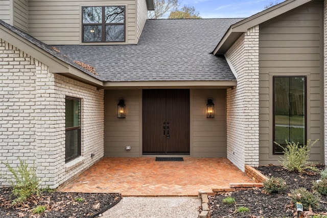 view of exterior entry featuring a patio, brick siding, and roof with shingles