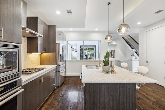 kitchen with pendant lighting, sink, stainless steel appliances, an island with sink, and wall chimney exhaust hood