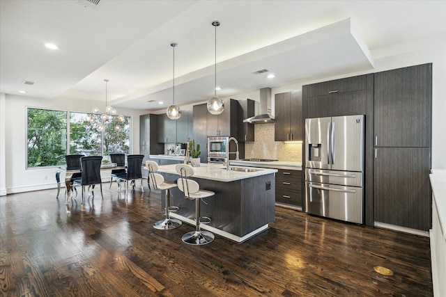 kitchen with wall chimney range hood, dark brown cabinets, a center island with sink, stainless steel fridge with ice dispenser, and decorative light fixtures