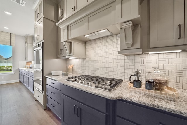 kitchen with appliances with stainless steel finishes, gray cabinetry, backsplash, light stone counters, and light wood-type flooring