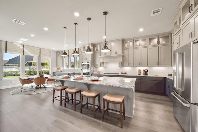 kitchen featuring a kitchen island with sink, stainless steel appliances, light stone countertops, decorative backsplash, and decorative light fixtures