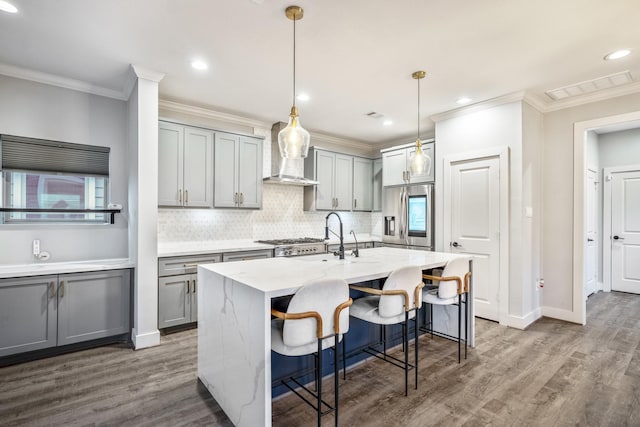 kitchen with gray cabinetry, stainless steel fridge, hanging light fixtures, a kitchen island with sink, and wall chimney exhaust hood