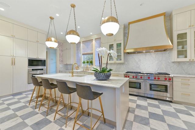 kitchen featuring premium range hood, light floors, an island with sink, stainless steel appliances, and a sink