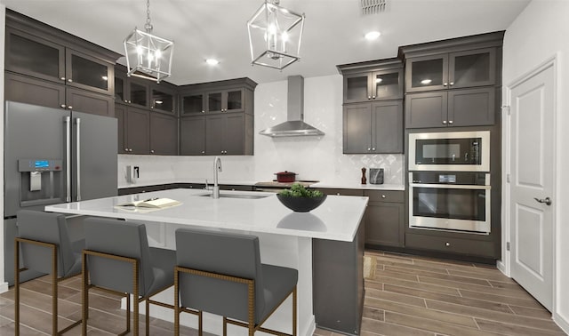 kitchen with wall chimney exhaust hood, sink, an inviting chandelier, an island with sink, and stainless steel appliances