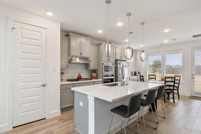 kitchen featuring a breakfast bar, decorative light fixtures, gray cabinetry, stainless steel appliances, and a center island with sink