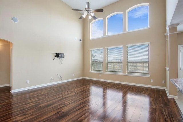 unfurnished living room with dark wood-style floors, arched walkways, a high ceiling, ceiling fan, and baseboards