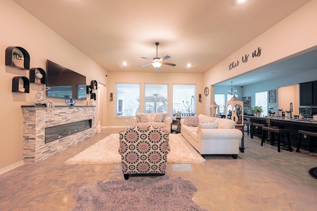 living room with concrete flooring, a stone fireplace, and ceiling fan with notable chandelier