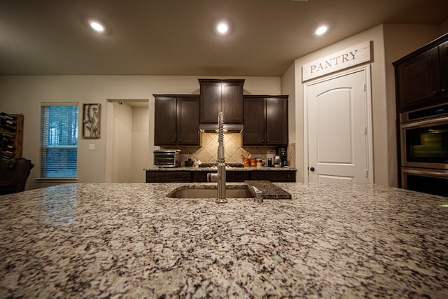 kitchen featuring sink, light stone counters, dark brown cabinets, double oven, and backsplash
