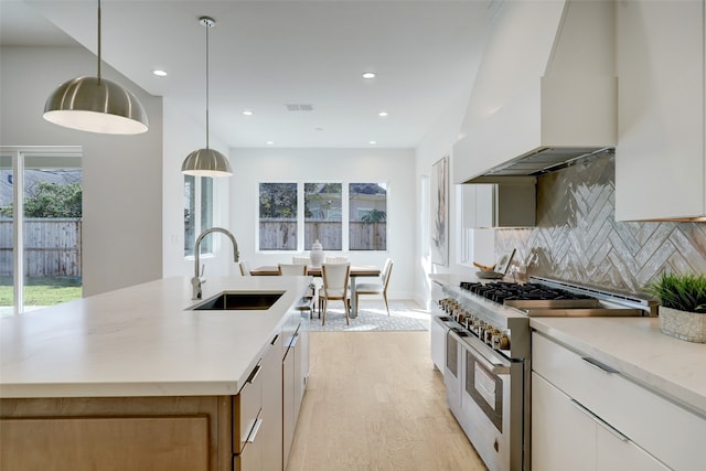 kitchen featuring custom exhaust hood, white cabinetry, a center island with sink, pendant lighting, and range with two ovens