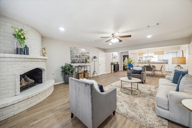 living room with a textured ceiling, ceiling fan with notable chandelier, a brick fireplace, and light hardwood / wood-style flooring