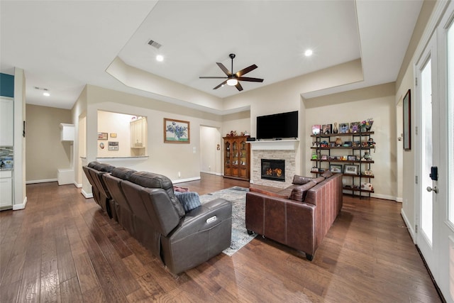 living room featuring a raised ceiling, ceiling fan, a stone fireplace, and dark hardwood / wood-style flooring