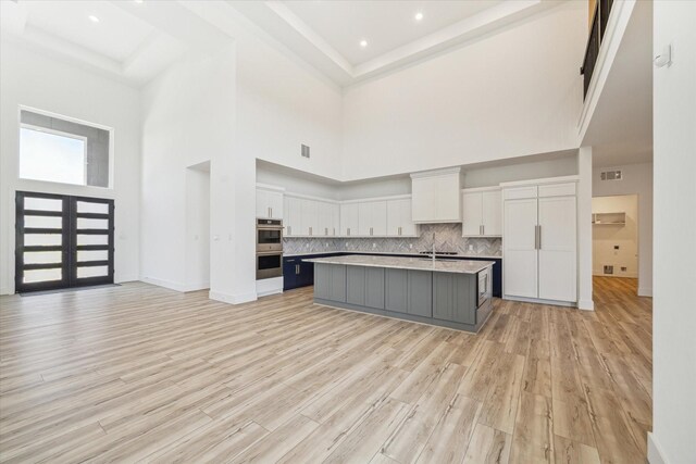 kitchen with white cabinetry, double oven, backsplash, light hardwood / wood-style floors, and a center island with sink