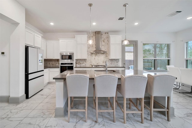 kitchen featuring pendant lighting, wall chimney range hood, appliances with stainless steel finishes, white cabinetry, and an island with sink