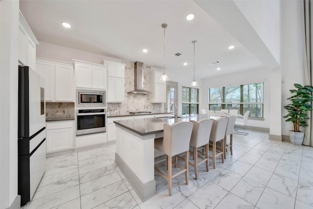 kitchen with wall chimney range hood, sink, a kitchen island with sink, stainless steel appliances, and white cabinets