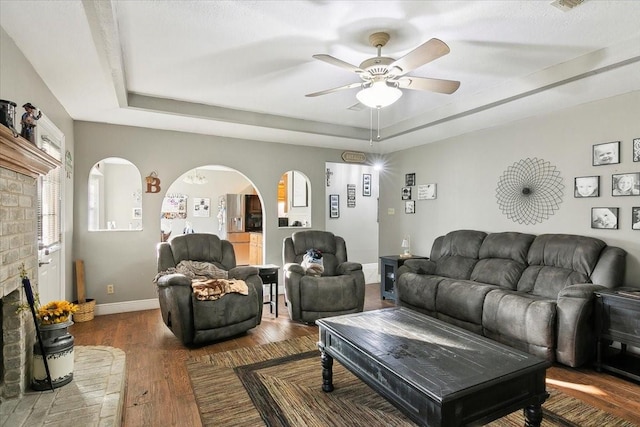 living room featuring dark wood-type flooring, ceiling fan, a raised ceiling, and a brick fireplace