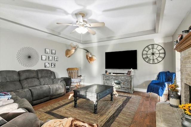 living room featuring ceiling fan, wood-type flooring, a raised ceiling, and a brick fireplace