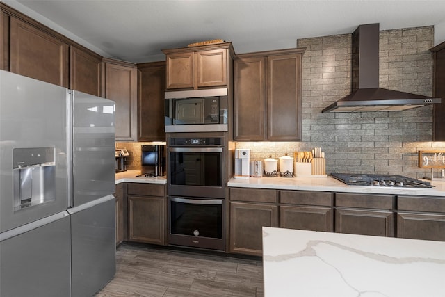 kitchen featuring stainless steel appliances, wall chimney range hood, light stone counters, and decorative backsplash