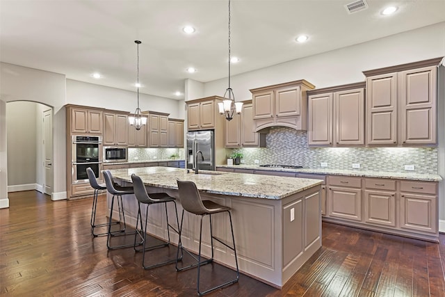 kitchen with an island with sink, appliances with stainless steel finishes, light stone counters, and decorative light fixtures