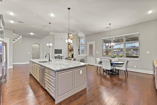 kitchen with sink, dark wood-type flooring, an inviting chandelier, light stone counters, and an island with sink