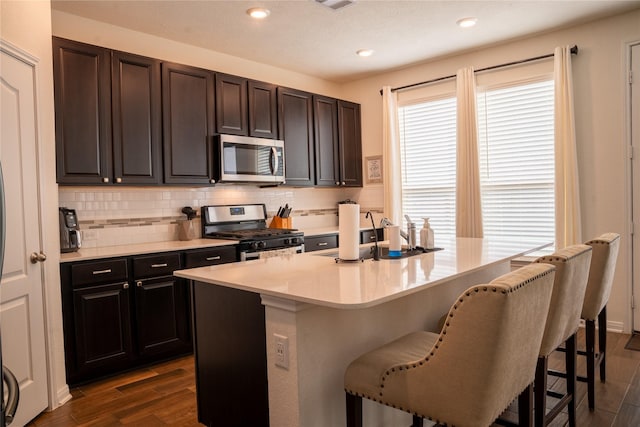 kitchen with dark wood-type flooring, a kitchen bar, tasteful backsplash, appliances with stainless steel finishes, and a kitchen island with sink