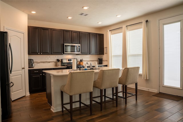 kitchen with a breakfast bar, an island with sink, dark hardwood / wood-style flooring, decorative backsplash, and stainless steel appliances