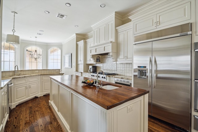 kitchen featuring butcher block counters, sink, hanging light fixtures, stainless steel built in fridge, and a center island with sink