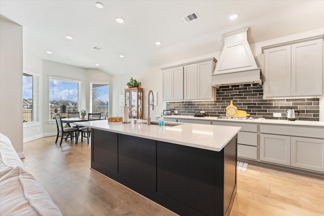 kitchen with gray cabinets, sink, backsplash, custom exhaust hood, and a kitchen island with sink