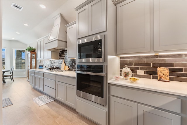 kitchen featuring lofted ceiling, gray cabinetry, backsplash, stainless steel appliances, and custom range hood