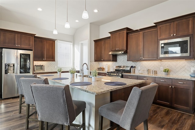 kitchen with sink, appliances with stainless steel finishes, hanging light fixtures, light stone counters, and an island with sink