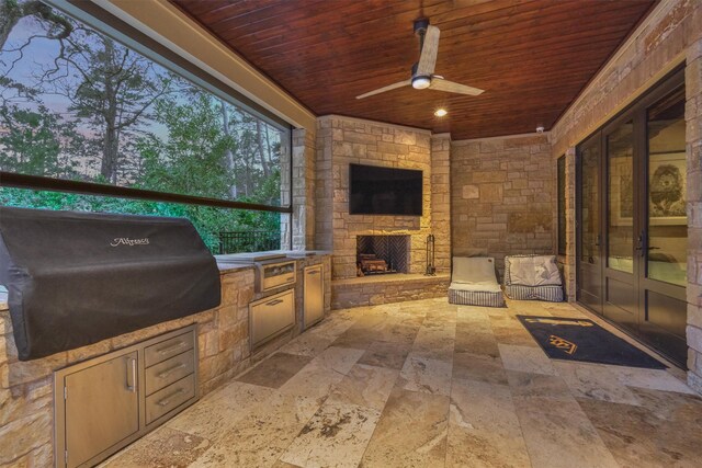 patio terrace at dusk featuring ceiling fan, area for grilling, and an outdoor stone fireplace