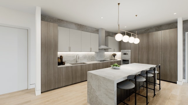 kitchen with wall chimney range hood, sink, white cabinetry, hanging light fixtures, and a center island