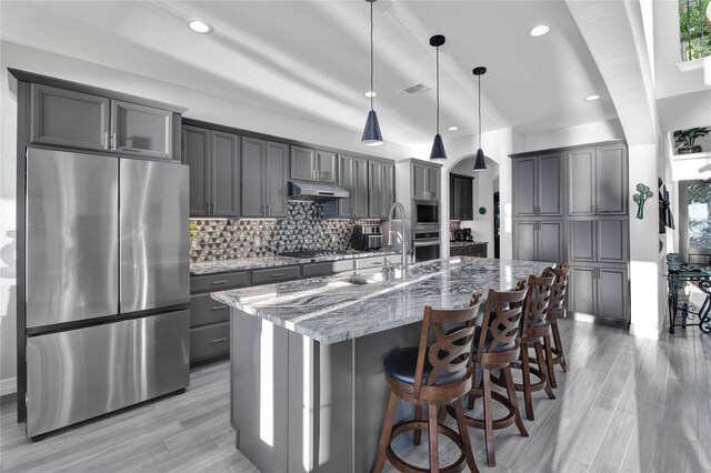 kitchen featuring appliances with stainless steel finishes, pendant lighting, backsplash, a kitchen island with sink, and light stone counters