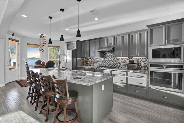 kitchen with tasteful backsplash, pendant lighting, stainless steel appliances, light stone countertops, and a kitchen island with sink