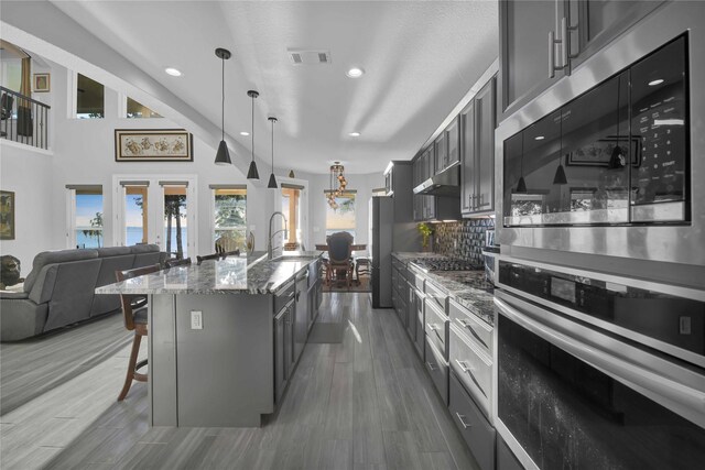 kitchen featuring dark stone countertops, hanging light fixtures, a center island with sink, stainless steel appliances, and a kitchen bar