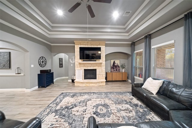 living room featuring crown molding, ceiling fan, a tray ceiling, wood-type flooring, and a stone fireplace