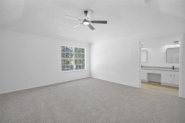 unfurnished bedroom with ensuite bathroom, built in desk, ceiling fan, light carpet, and a textured ceiling