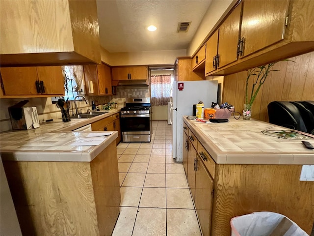 kitchen featuring light tile patterned flooring, stainless steel gas stove, sink, white fridge, and tile counters